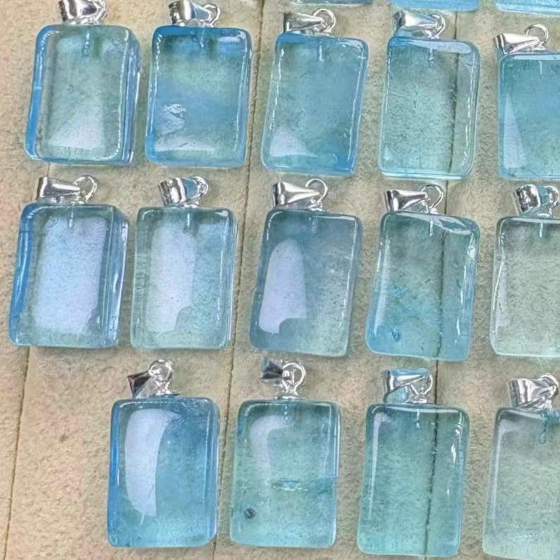 May include: A collection of rectangular, light blue pendants with silver-colored bails. The pendants have a translucent, glassy appearance, and the color varies slightly between each piece. The pendants are arranged closely together, suggesting they are for sale.
