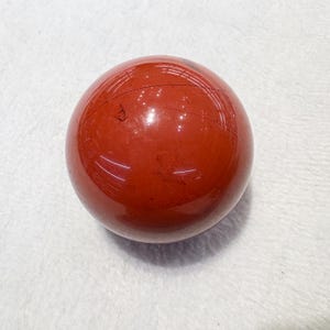 May include: A polished, spherical gemstone in a deep red hue. The orb has a smooth, reflective surface with subtle natural markings. The gemstone is displayed on a white surface. This item is suitable for home decor or as a collectible.