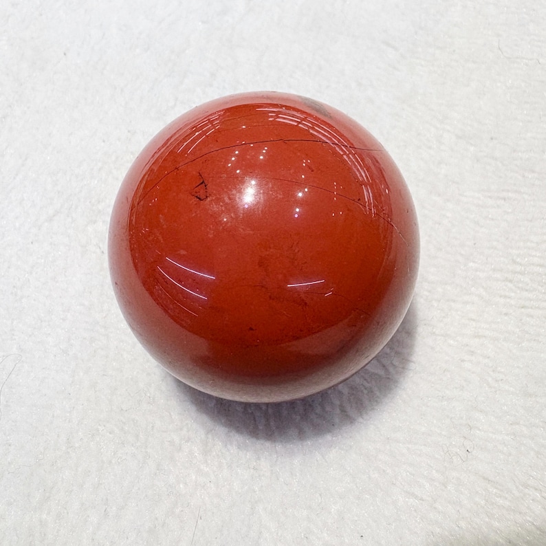 May include: A polished, spherical gemstone in a deep red hue. The orb has a smooth, reflective surface with subtle natural markings. The gemstone is displayed on a white surface. This item is suitable for home decor or as a collectible.