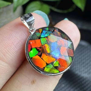 May include: Round pendant with a silver bail, featuring a mosaic of vibrant, iridescent colors including red, orange, green, and blue. The gemstone is set in a silver bezel, creating a striking contrast. The pendant is a unique piece of jewelry.
