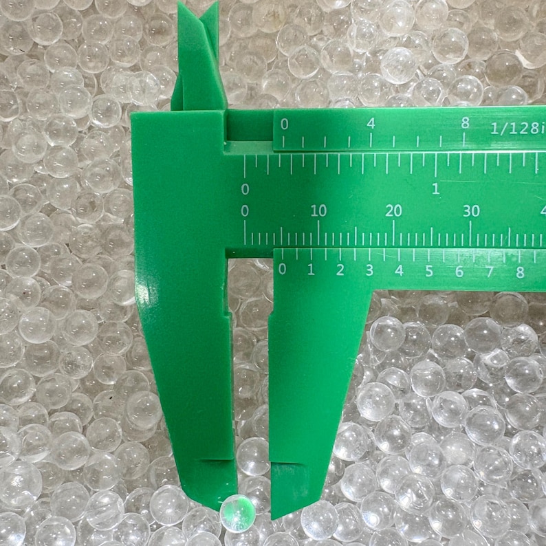 May include: A close-up of a green caliper measuring a small, iridescent sphere. The caliper shows measurements in inches, with the sphere appearing to be less than 1/8 inch in diameter. The background is filled with many clear spheres.