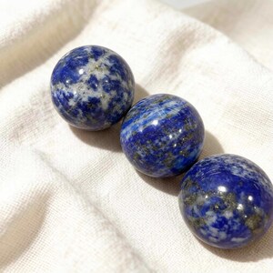 May include: Three polished lapis lazuli spheres. The spheres are a deep blue colour with white and gold flecks. The spheres are arranged on a white cloth.