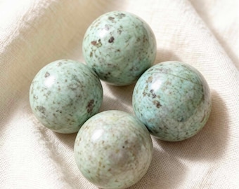 May include: Four spherical, speckled, light green stones arranged on a cream-colored fabric. The stones have a polished appearance with darker green and brown spots. The spheres are approximately 2 inches in diameter, and are likely decorative or collectible items.