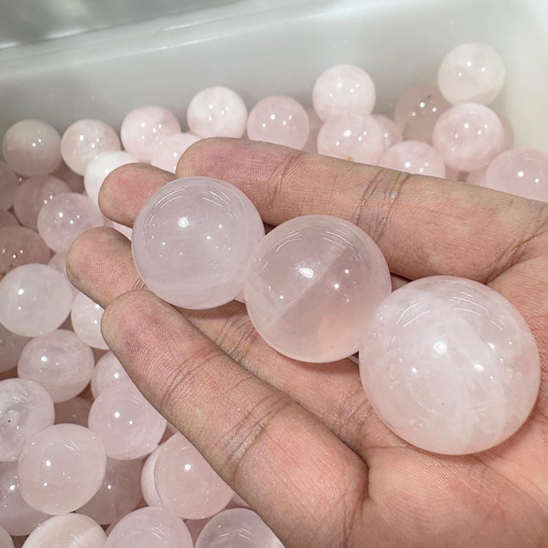 May include: A collection of translucent, pale pink rose quartz spheres. Three spheres are held in a hand, with many more visible in the background. The spheres have a smooth, polished appearance, and the color is consistent throughout.
