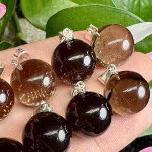 May include: A selection of round, polished gemstone pendants in shades of brown and black. Each pendant has a silver-coloured bail. The gemstones are translucent, with a visible internal pattern. The pendants are displayed against a soft pink and green background.