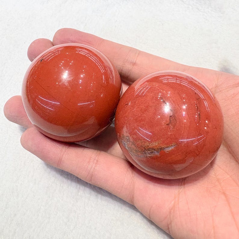 May include: Two polished red jasper spheres. The spheres are a deep reddish-brown color with subtle variations in tone and veining. The smooth, rounded stones are held in a hand, showcasing their size and texture. The spheres are a natural stone product.