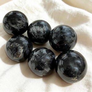 Può includere: Sei sfere nere lucide con inclusioni bianche e grigie sono disposte su un tessuto bianco. Le sfere hanno una finitura lucida, che riflette la luce. L'estetica generale è naturale ed elegante, suggerendo una collezione di pietre decorative o cristalli.