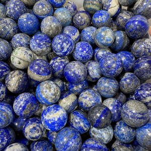 May include: A close-up of numerous polished lapis lazuli spheres. The stones are a rich blue with grey and white veining. The spheres are of varying sizes and are closely arranged, giving a textured look.