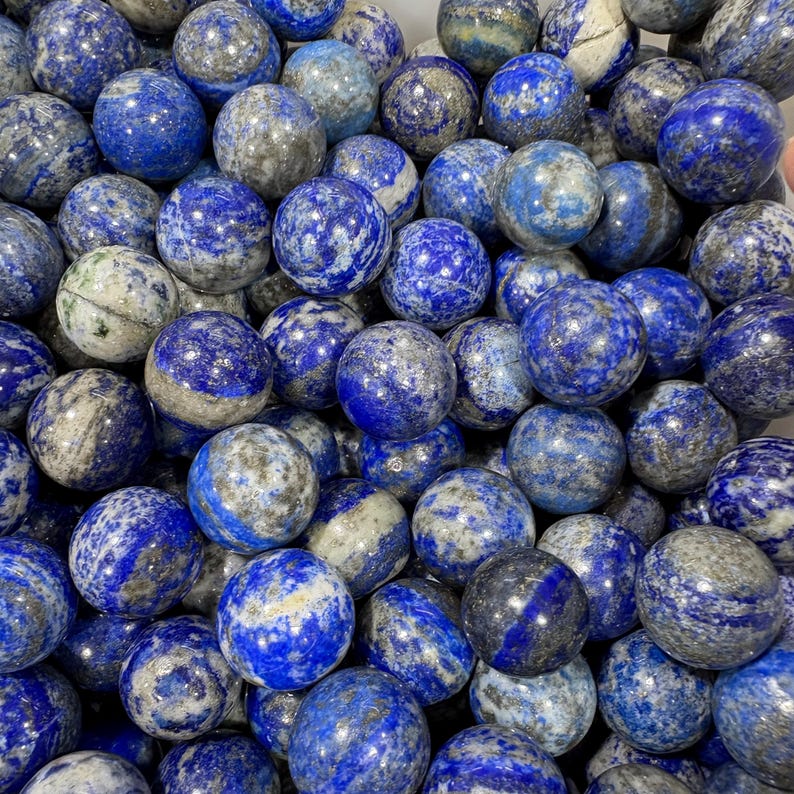 May include: A close-up of numerous polished lapis lazuli spheres. The stones are a rich blue with grey and white veining. The spheres are of varying sizes and are closely arranged, giving a textured look.
