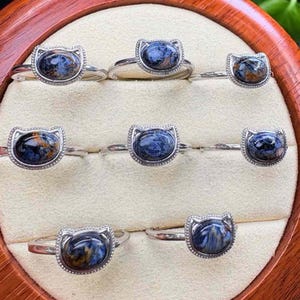 May include: A collection of silver rings, each featuring a cat-shaped setting. The rings showcase oval-shaped gemstones with a mix of blue, brown, and gold hues. The rings are displayed in a wooden jewelry box with a cream-colored interior.