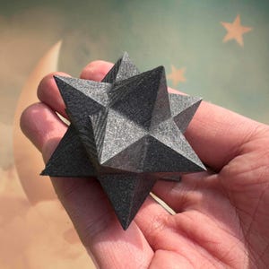 Peut inclure: Un objet géométrique en forme d'étoile, de couleur gris foncé, avec des bords pointus. L'objet est tenu dans une main, mettant en valeur son design complexe et sa surface texturée. L'arrière-plan présente un thème céleste avec une lune et des étoiles.