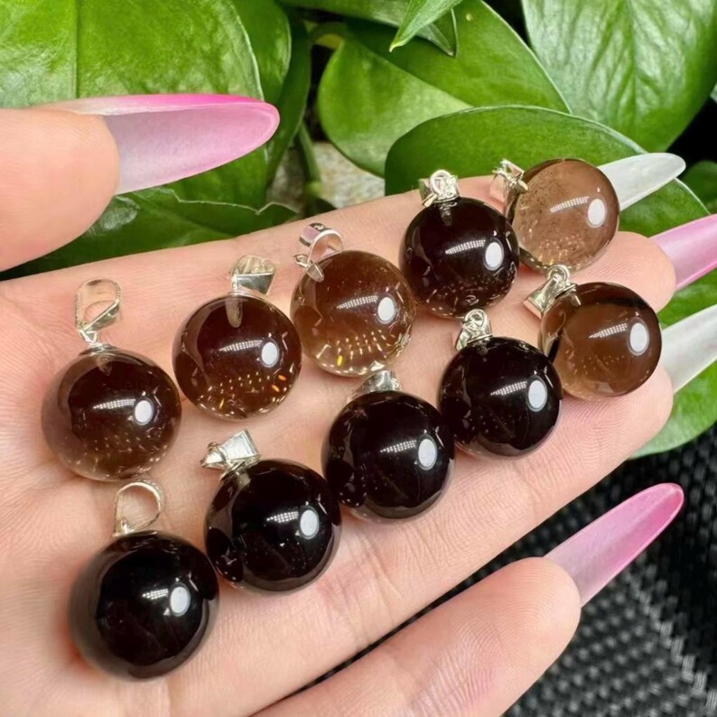 May include: A selection of spherical gemstone pendants in shades of brown and black. Each pendant has a silver-coloured bail for attaching to a chain. The gemstones are smooth and reflective, with a glossy finish.
