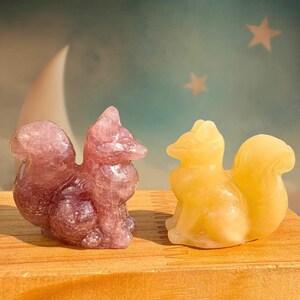 Könnte beinhalten: Zwei geschnitzte Eichhörnchenfiguren. Eine ist tiefviolett, die andere hellgelb. Die Eichhörnchen sitzen auf einer Holzoberfläche, mit einem verträumten Hintergrund aus Mond und Sternen.