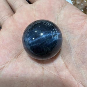 以下が含まれることがあります： 光沢のある濃い青色の球体で、虹色の薄い青色の縞模様があります。 宝石のボールは手で持たれており、その滑らかで反射する表面を示しています。 球体の色と模様は、魅力的な視覚効果を生み出しています。
