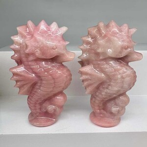 May include: Two pink seahorse sculptures. Each seahorse is carved from a translucent, light pink stone. The sculptures have detailed fins, spiky dorsal fins, and rounded bodies. They are displayed on a white surface.