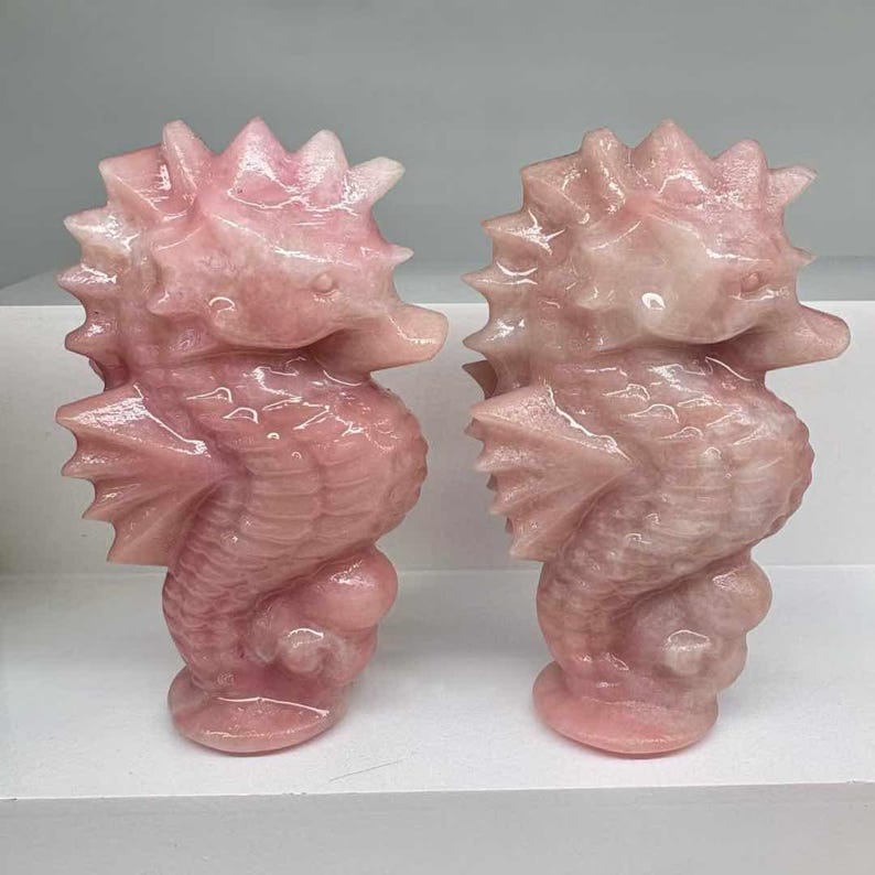 May include: Two pink seahorse sculptures. Each seahorse is carved from a translucent, light pink stone. The sculptures have detailed fins, spiky dorsal fins, and rounded bodies. They are displayed on a white surface.