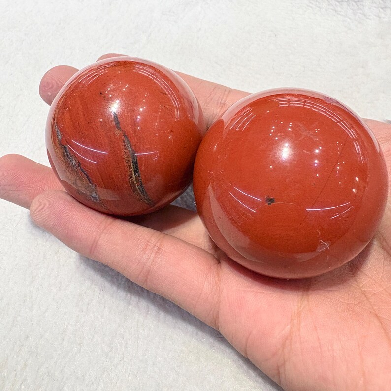 May include: Two polished red jasper spheres, each held in a hand. The spheres have a smooth, reflective surface with natural veining and markings. The color is a deep, rich red with variations in tone.