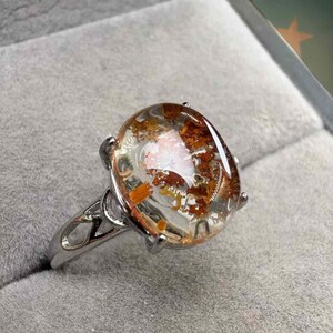 May include: A silver ring featuring a large, oval-shaped gemstone. The clear stone has inclusions of orange and brown, creating a unique pattern. The ring's band has an open, decorative design.