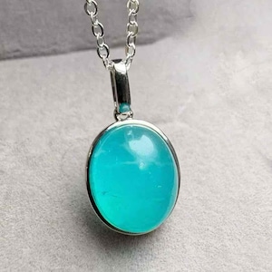 May include: A silver necklace with a pendant showcasing a bright turquoise oval stone. The stone is set in a silver bezel and suspended from a fine chain. The pendant's vivid colour provides a striking contrast against the silver chain.
