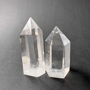 May include: Two clear quartz crystal points with hexagonal facets and pointed terminations. The taller crystal stands to the left, while the slightly shorter, wider crystal is positioned to the right, both displaying natural internal inclusions.