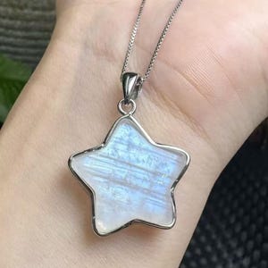 May include: A silver necklace with a star-shaped pendant. The pendant features a light blue and white iridescent stone set in a silver frame. The necklace is shown against a neutral background.