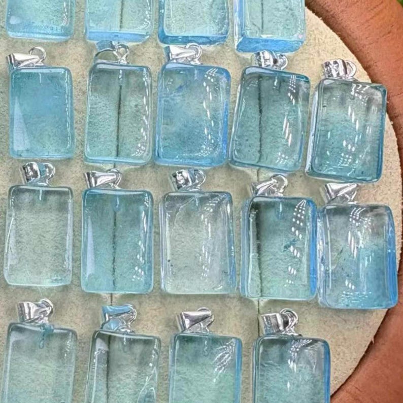 May include: A collection of rectangular, light blue gemstone pendants with silver-colored bails. The pendants are arranged closely together, showcasing their translucent quality and varying shades of blue. The overall aesthetic is one of delicate jewelry.