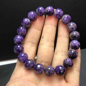 Puede incluir: Un brazalete de cuentas redondas y pulidas de charoíta púrpura. El brazalete se sostiene en una mano, mostrando las diferentes tonalidades de púrpura y los patrones naturales dentro de cada cuenta. Las cuentas tienen aproximadamente 1,5 cm de diámetro.