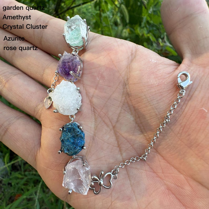 May include: A silver bracelet featuring a series of colorful raw gemstones, including garden quartz, amethyst, crystal cluster, azurite, and rose quartz. The bracelet has a butterfly charm and a chain extender.