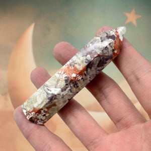 May include: A multi-colored crystal point held in a hand. The crystal features a mix of white, gray, red, and green hues. The crystal is approximately 4 inches long and has a polished, smooth surface. The background is a soft blend of colors.