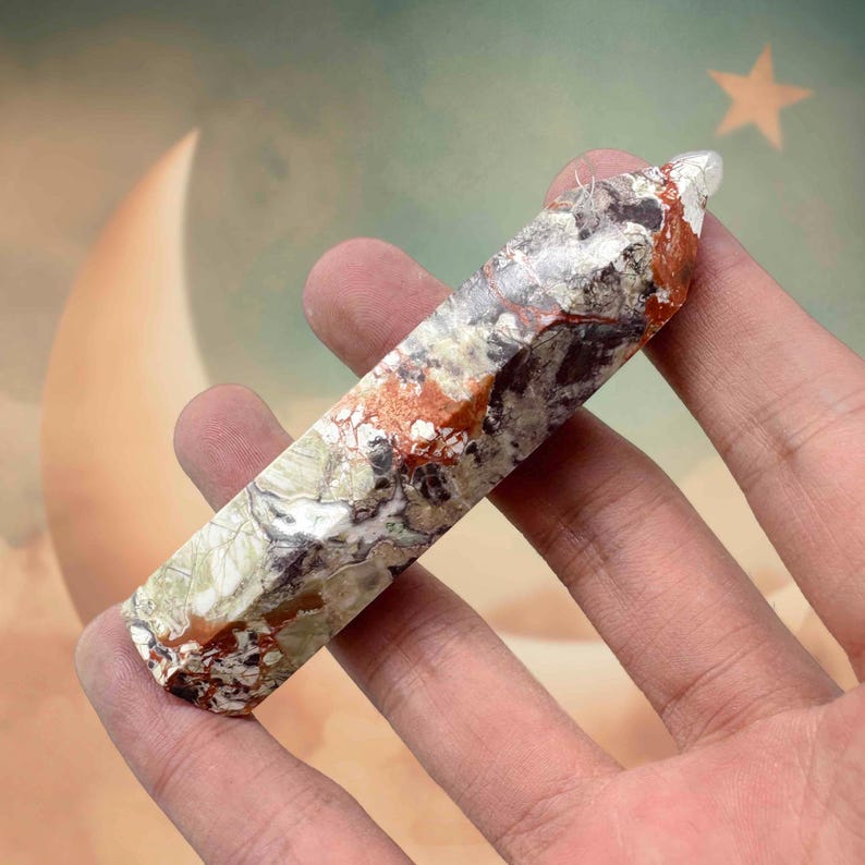 May include: A multi-colored crystal point held in a hand. The crystal features a mix of white, gray, red, and green hues. The crystal is approximately 4 inches long and has a polished, smooth surface. The background is a soft blend of colors.