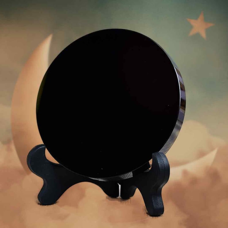 May include: A large, circular, polished black crystal is displayed on a small black wooden stand. The crystal is the main focus, reflecting light. The background has a soft, dreamy scene with a crescent moon and a star.