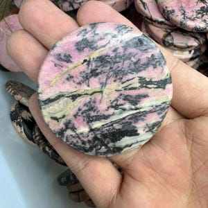 May include: A round, polished stone with a pink base color, veined with black, white, and yellow patterns. The stone is held in a hand, with other similar stones visible in the background. The stone is a decorative item.