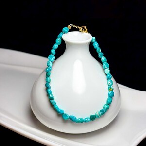May include: A bracelet composed of small, irregular, tumbled turquoise beads in shades of bright blue and teal. The beads are strung together and secured with a gold-toned metal clasp mechanism at the top, featuring a small circular ring and hook.