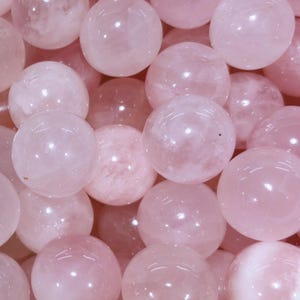 May include: A close-up shot of multiple rose quartz spheres. The spheres are a soft pink color, with a smooth, polished surface. The spheres are of varying sizes and are clustered together, filling the frame.