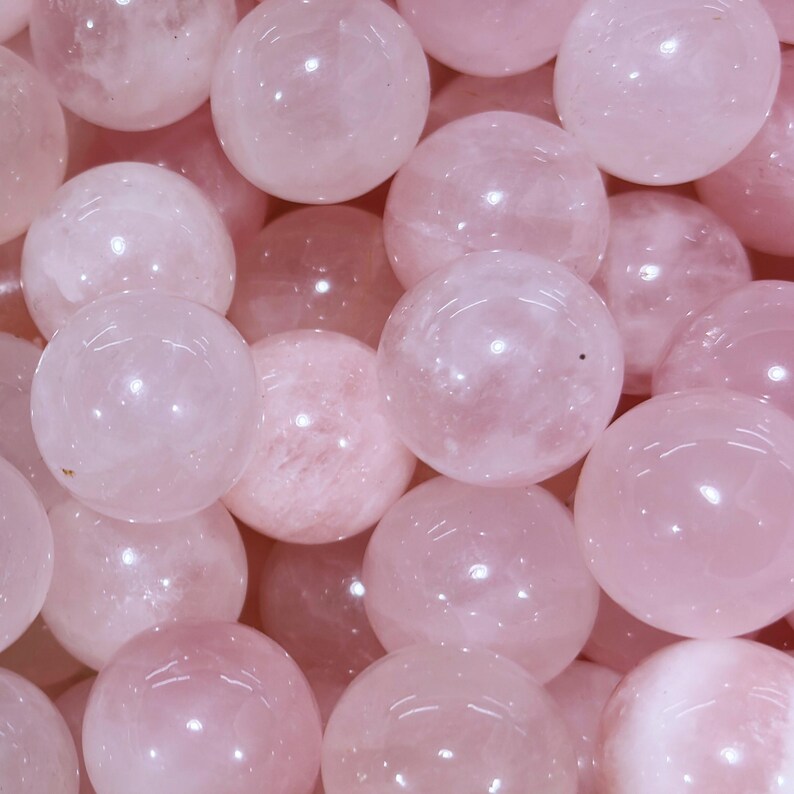 May include: A close-up shot of multiple rose quartz spheres. The spheres are a soft pink color, with a smooth, polished surface. The spheres are of varying sizes and are clustered together, filling the frame.