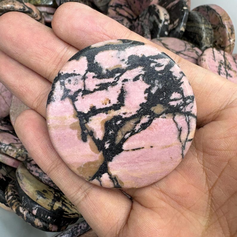 May include: A round, polished gemstone with a pink base and black veining. The stone is held in a hand, showcasing its smooth surface and natural patterns. Other similar stones are visible in the background.
