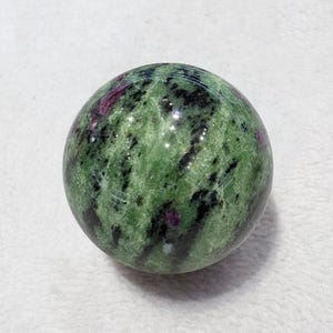 May include: A polished sphere of green zoisite with ruby inclusions. The stone has a mottled appearance with dark green and black patches, and small flecks of red ruby. The sphere is smooth and round, and appears to be made of natural stone.