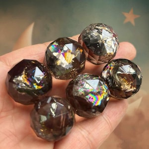 Può includere: Una collezione di sfere sfaccettate di quarzo fumé. Le sfere hanno un aspetto scuro e traslucido con iridescenza interna simile all'arcobaleno. Il taglio delle sfere crea molteplici superfici riflettenti, esaltando il loro fascino visivo. Le sfere sono una gemma naturale.
