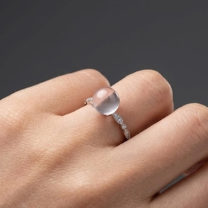May include: A silver-toned ring featuring a large, smooth, translucent, pale pink spherical gemstone set atop a slender band. The band is decorated with a series of small, clear, sparkling stones arranged in a delicate, elongated marquise-like pattern.