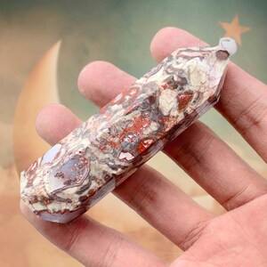 May include: A polished, multi-colored gemstone wand. The wand features a mix of red, white, and gray patterns. The top of the wand has a small, clear, rounded piece. The gemstone is held in a hand.