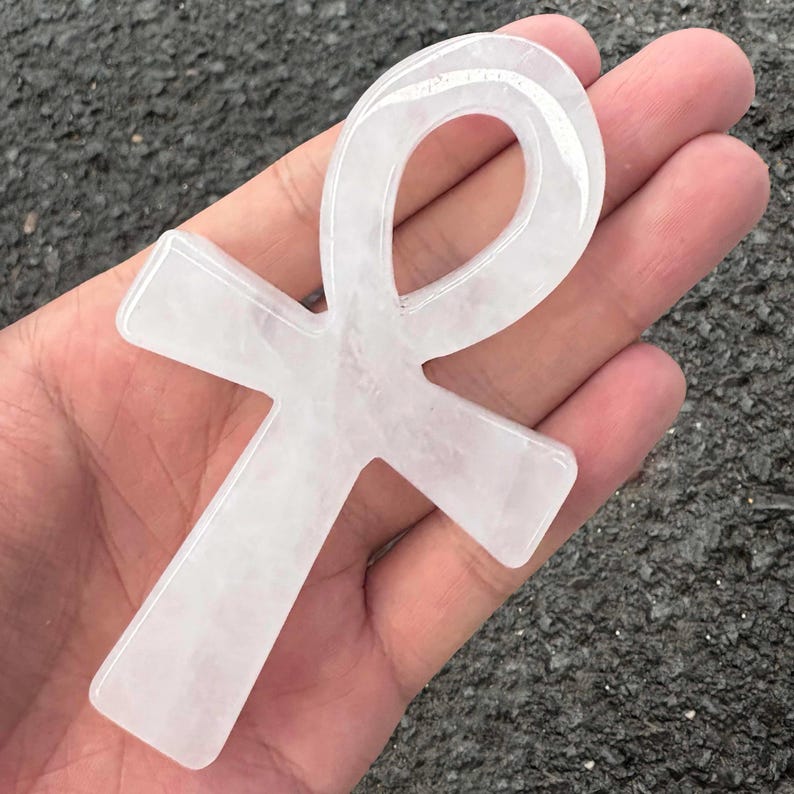 May include: A polished, translucent white quartz crystal carved into the shape of an Egyptian ankh symbol. The stone features a smooth, glossy surface with subtle internal cloudy inclusions and a distinct cross-like form with a looped top.