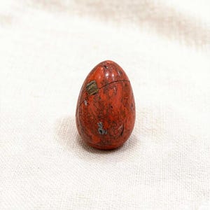 Peut inclure: Une pierre polie en forme d'œuf avec un motif rouge et brun moucheté. La pierre présente quelques petites fissures et un petit patch rectangulaire d'un matériau différent. L'œuf est posé debout sur une surface blanche.