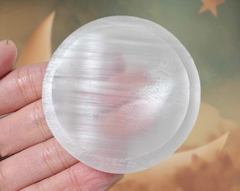 May include: A round, translucent selenite crystal plate. The plate is held in a hand, showcasing its smooth, polished surface. The crystal has a subtle, natural texture and a soft, white color. The background features a celestial-themed design.