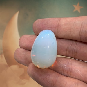 May include: A polished, egg-shaped gemstone with a pearly, opalescent appearance. The stone has a smooth surface with a gradient of colors, transitioning from a soft blue at the top to a warm, orange hue at the base. The gemstone is held in a hand.