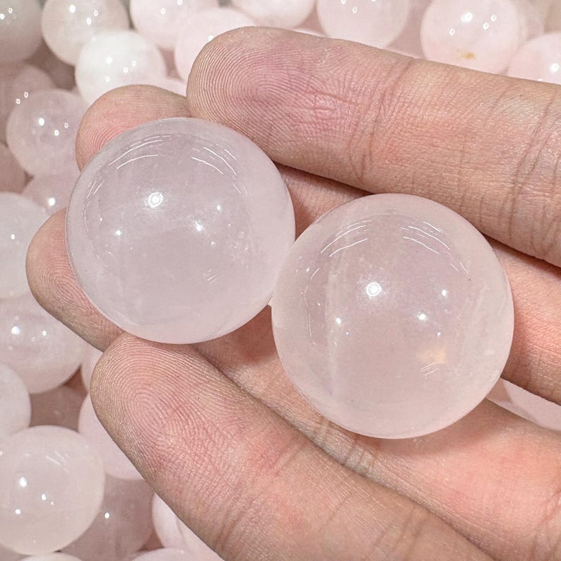 May include: Two translucent, pale pink rose quartz spheres are held in a hand, with several more spheres visible in the background. The spheres have a smooth, polished surface and a soft, glowing appearance. These spheres are a popular choice for crystal healing and meditation.