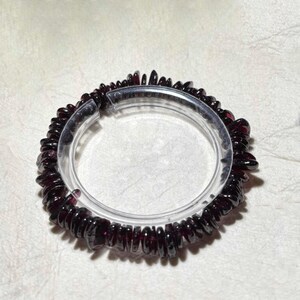 Puede incluir: Una pulsera circular compuesta por numerosas pequeñas esquirlas irregulares de granate de color rojo intenso ensartadas. La pulsera se muestra sobre un soporte circular de plástico transparente, resaltando la naturaleza oscura, translúcida y pulida de las piedras.
