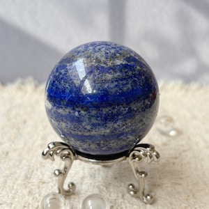 Puede incluir: Una esfera pulida de lapislázuli, que muestra un tono azul profundo con vetas grises y motas doradas. La bola de piedra preciosa descansa sobre un soporte de metal plateado ornamentado. La esfera es un objeto decorativo.