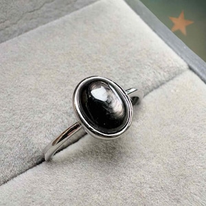 Puede incluir: Un anillo de plata con una gran piedra preciosa negra de forma ovalada. La piedra preciosa está engastada en un bisel de plata y tiene una superficie reflectante. El anillo se muestra sobre una superficie gris.