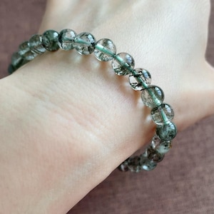 May include: A bracelet featuring clear, round beads with green inclusions. The beads are strung together on a thin, green elastic cord. The bracelet is shown on a person's wrist.