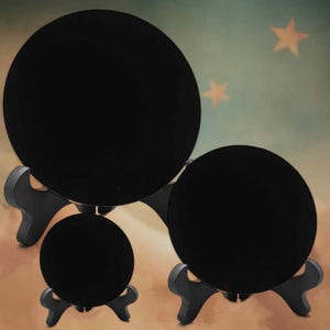 May include: Three black, circular display stands of different sizes. Each stand supports a round object, forming a tiered arrangement. The stands are crafted from dark wood, designed for displaying items. The background has a soft gradient with star shapes.